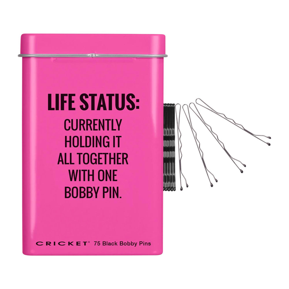 A bright pink rectangular tin with a lid, featuring black text that reads 'LIFE STATUS: CURRENTLY HOLDING IT ALL TOGETHER WITH ONE BOBBY PIN.' and 'CRICKET 75 Black Bobby Pins' at the bottom, shown on a plain white background.