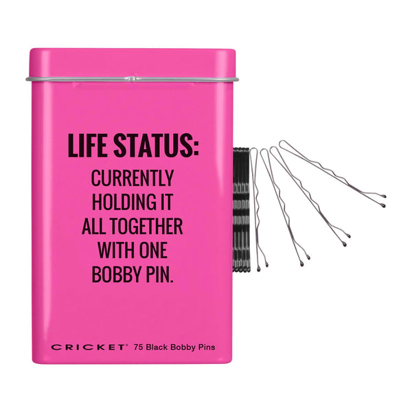 A bright pink rectangular tin labeled with humorous text about holding it together with one bobby pin, shown with a spool of black bobby pins and several loose pins on a white background.