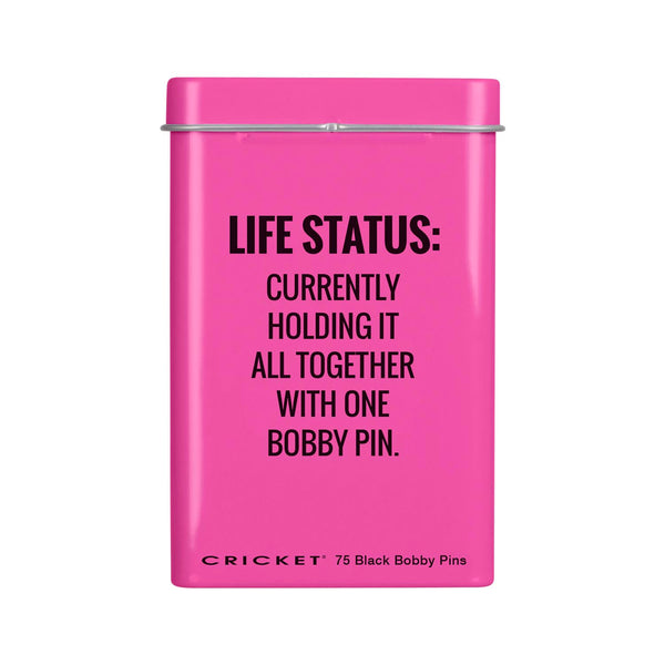A bright pink rectangular tin with a lid, featuring black text that reads 'LIFE STATUS: CURRENTLY HOLDING IT ALL TOGETHER WITH ONE BOBBY PIN.' and 'CRICKET 75 Black Bobby Pins' at the bottom, shown on a plain white background.