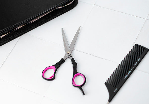 Angled view of open Style Xpress La-Di-Da 5.75" Shear, handle is black with hot pink accents on tile next to the Carbon Rattail Comb and a zipper case