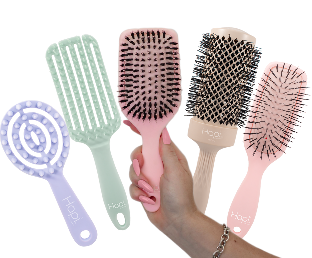 five Hapi hair care tools are shown on a white background: a lavender spiral detangling brush, a mint green wide-tooth comb, a pink paddle brush being held in a hand, and a pink round brush. A hand with pink nail polish is visible holding the pink paddle brush.