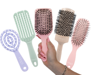 five Hapi hair care tools are shown on a white background: a lavender spiral detangling brush, a mint green wide-tooth comb, a pink paddle brush being held in a hand, and a pink round brush. A hand with pink nail polish is visible holding the pink paddle brush.