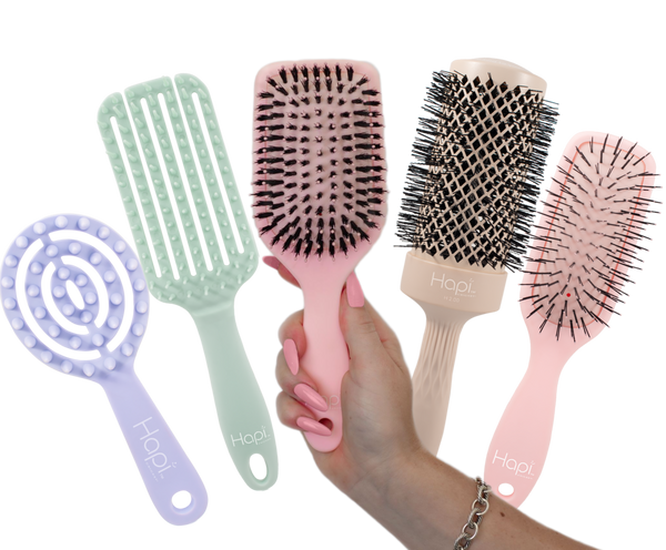 five Hapi hair care tools are shown on a white background: a lavender spiral detangling brush, a mint green wide-tooth comb, a pink paddle brush being held in a hand, and a pink round brush. A hand with pink nail polish is visible holding the pink paddle brush.